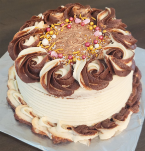 Beautifully decorated chocolate cake with swirls of chocolate and vanilla frosting, topped with colorful sprinkles and edible pearls, created by Cake Coma