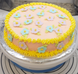 Delightful flower-themed cake decorated with pastel-colored fondant flowers and yellow frosting, created by Cake Coma.